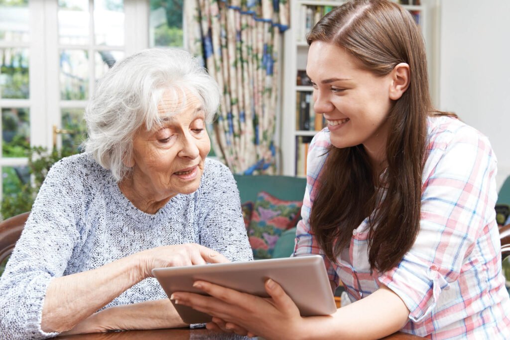 A young woman helps an elderly woman use a tablet device at a table. They are both smiling, and natural light comes in through the window behind them. The setting appears to be a cozy, well-lit home.