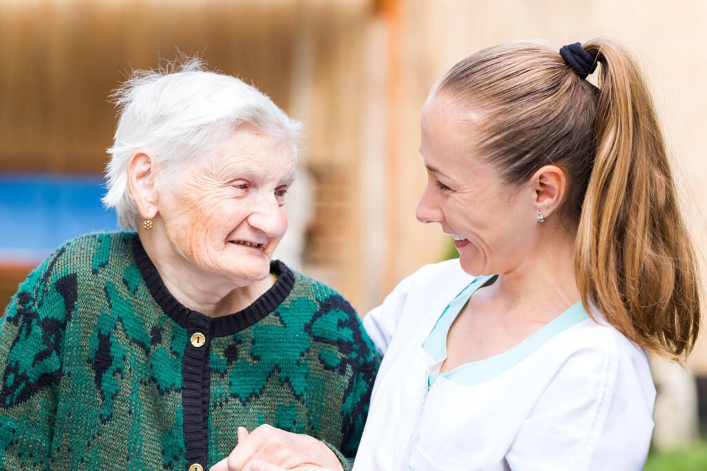 A smiling elderly woman with white hair wearing a green sweater holds hands and shares a joyful moment with a young female caregiver in a white uniform, both looking at each other warmly outdoors.