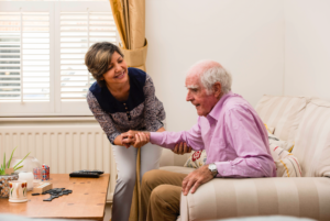 A smiling woman helps an elderly man stand up from a sofa in a bright living room holding his hand supportively A coffee table with mugs and dominoes is in the foreground